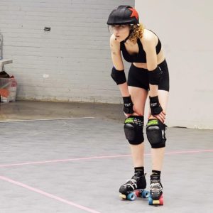 Tall white woman in black roller derby gear: skates, helmet, elblow pads, knee pads